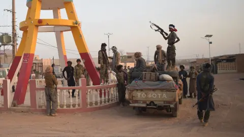 AFP via Getty Images A group of fighters in camouglage stand around a pick-up truck on a dusty road. One man in near silhouette - standing on the vehicle - is holding an automatic rifle up. On the left of the picture is the brown and yellow legs of a sculpture.