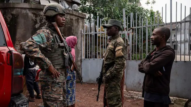 Deux hommes en uniforme militaire, fusils à la main, se tiennent aux côtés d'un passant vêtu d'un sweat-shirt à capuche marron et d'un pantalon. Une femme en sweat-shirt à capuche rose est de dos à la caméra, en arrière-plan.